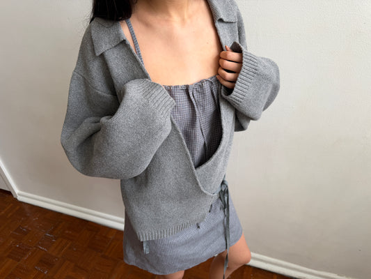 A cropped view of a woman wearing the Wrap Cardigan - Grey by Deiji Studios, a grey knit wrap cardigan with a collar, open over a grey top and mini skirt.