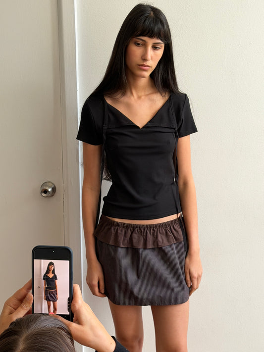 A woman with dark hair and a fringe stands against a door wearing the Flounce Layer Mini Skirt - Brown Blue Stripe by Deiji Studios, a fine brown-blue stripe mini skirt with a brown flounce waist, and a black tee, as another person photographs her on a phone.