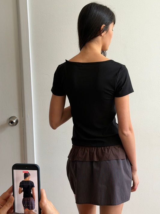 A woman with dark hair and a fringe stands with her back to the camera wearing the Flounce Layer Mini Skirt - Brown Blue Stripe by Deiji Studios, a fine brown-blue stripe mini skirt with a brown flounce waist, and a black tee.