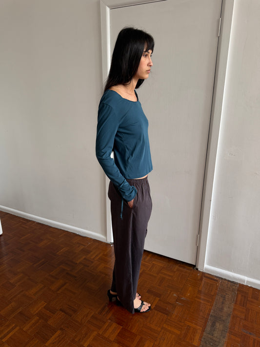 A woman with dark hair and a fringe stands in profile in a room wearing the Ease Trouser - Brown Blue Stripe by Deiji Studios, dark brown wide-leg trousers with a fine blue pinstripe, with a teal long-sleeve top and black sandals.
