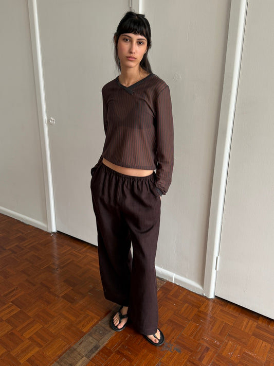 A woman with dark hair and a fringe stands in a room with hands in her pockets, wearing the Linen Ease Trouser - Brown Black Stripe by Deiji Studios, dark brown fine-striped wide-leg linen trousers, with a matching brown striped knit top and black flip-flops.