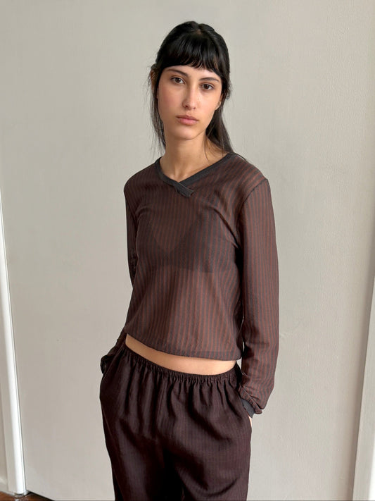 A woman with long dark hair and bangs wears the Stripe Longsleeve in Brown Grey Stripe, paired with matching pants, standing against a plain light-colored wall with her hands in her pockets.