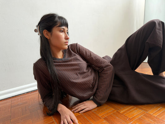 A person with long dark hair, wearing the Stripe Longsleeve in Brown Grey Stripe from Deiji Studios, lies on a wooden floor propped up on one arm, gazing thoughtfully to the side against a plain white wall.
