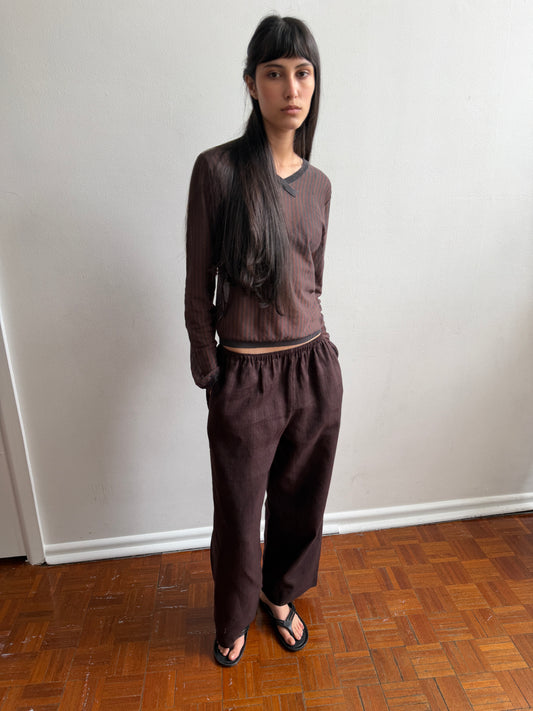 A woman with dark hair and a fringe stands in a room wearing the Linen Ease Trouser - Brown Black Stripe by Deiji Studios, dark brown fine-striped wide-leg linen trousers, with a matching brown striped knit top and black flip-flops.