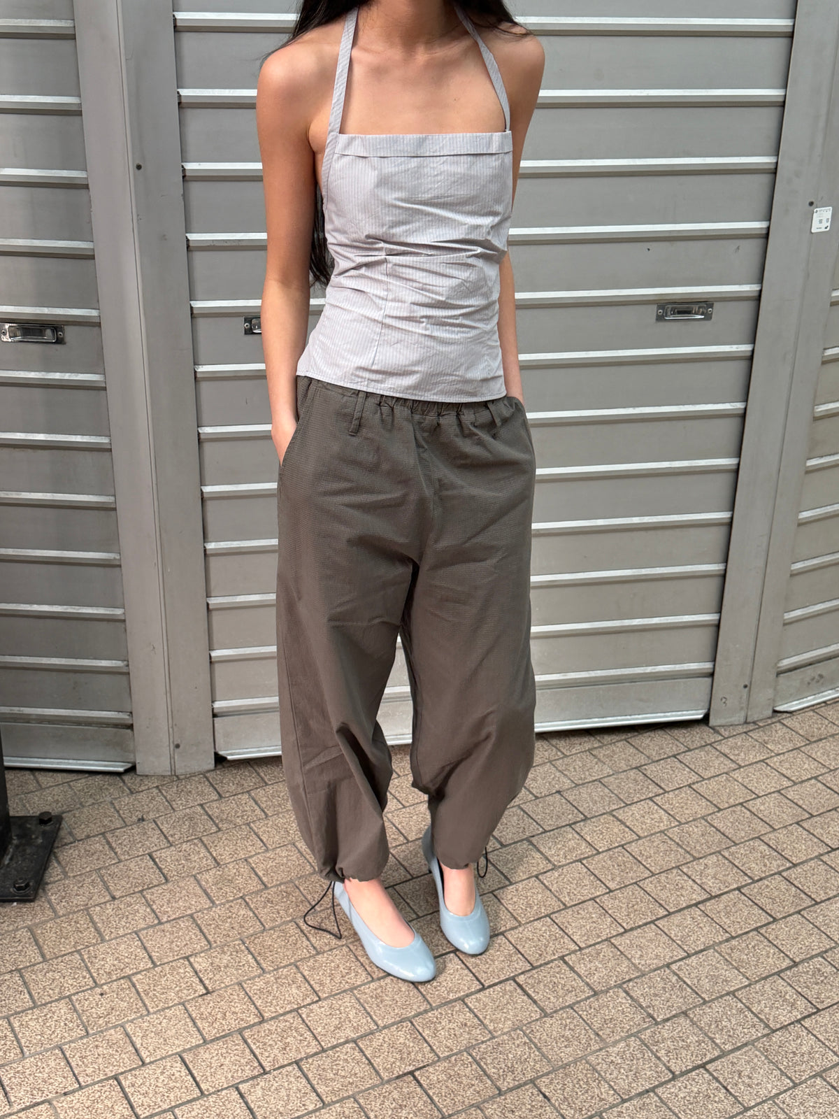 A cropped view of a woman outdoors wearing the Folded Top - Cloud Stripe by Deiji Studios, a pale grey-blue fine-striped fold-over halter top, with khaki trousers and pale blue heels.