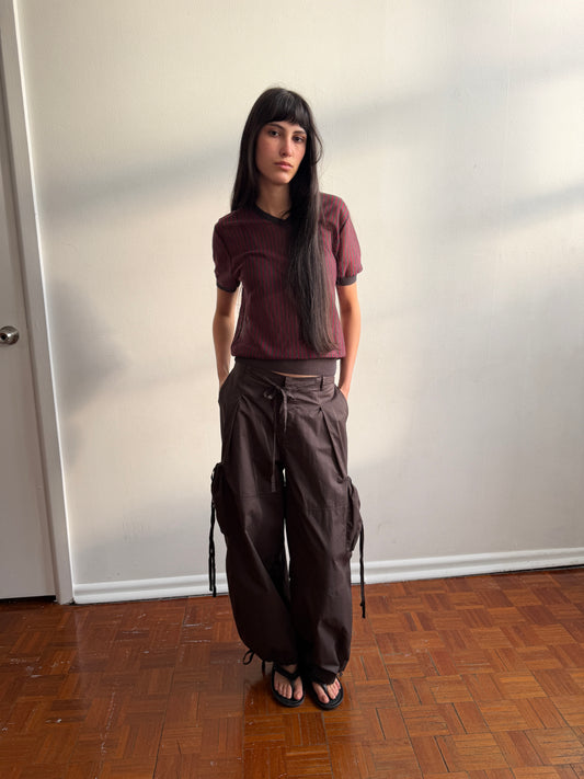 A woman with dark hair and a fringe stands against a wall in a room, wearing the Cross Neck Tee - Red Stripe by Deiji Studios, a fine red and charcoal striped knit tee, with dark brown wide-leg trousers and black sandals.