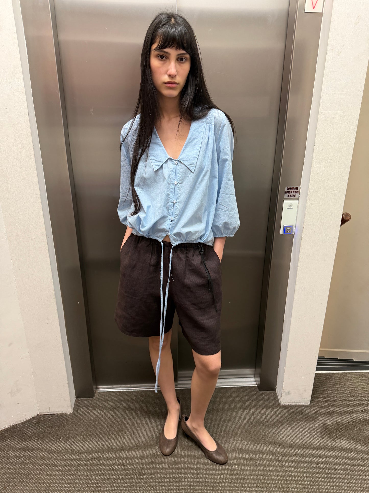 A woman with long dark hair stands in front of an elevator, wearing a light blue blouse and the Half Short in Brown Black Stripe, which features an elasticated waistband and wide leg opening, paired with brown flats. Her hands are in her pockets and she has a neutral expression.