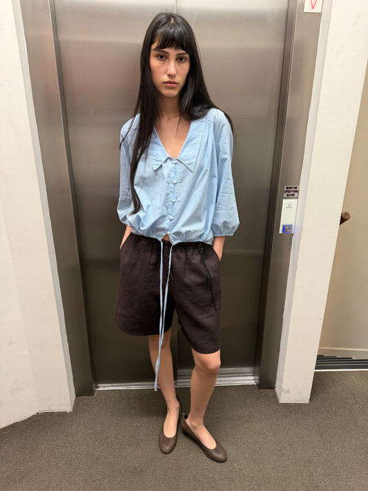 A woman with dark hair and a fringe stands in a lift wearing the Half Short - Brown Black Stripe by Deiji Studios, knee-length dark brown striped linen shorts, with a pale blue collared top and brown flats.