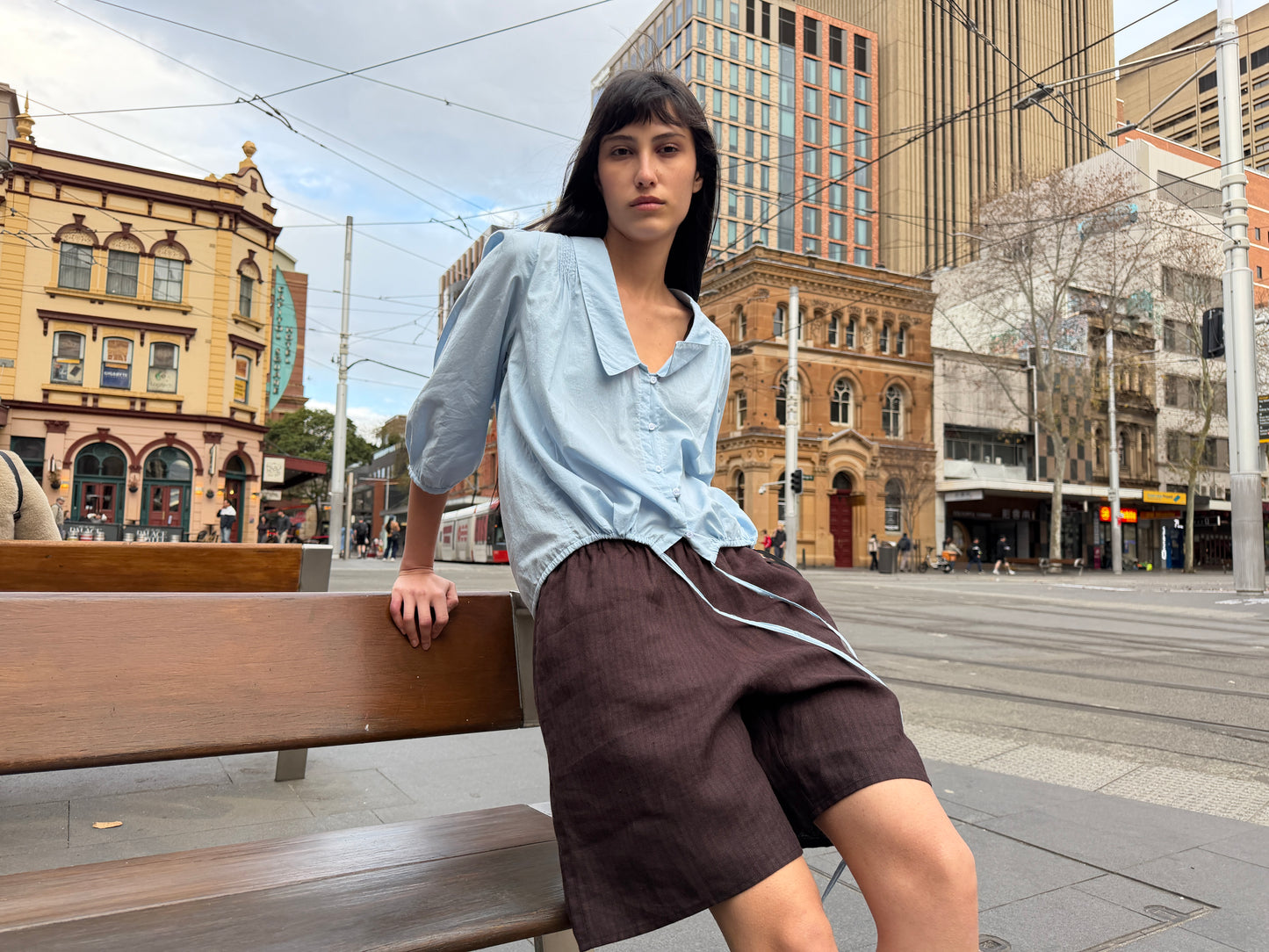 A person with long dark hair sits on a bench in a city square, wearing a light blue shirt and the Half Short - Brown Black Stripe, featuring relaxed linen fabric and an elasticated waistband. Historic and modern buildings, tram tracks, and overhead wires appear in the background.