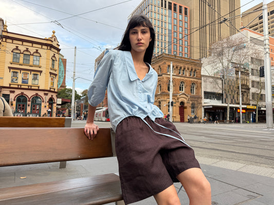 A woman with dark hair and a fringe sits on a bench in a city square wearing the Half Short - Brown Black Stripe by Deiji Studios, knee-length dark brown striped linen shorts, with a pale blue collared top.