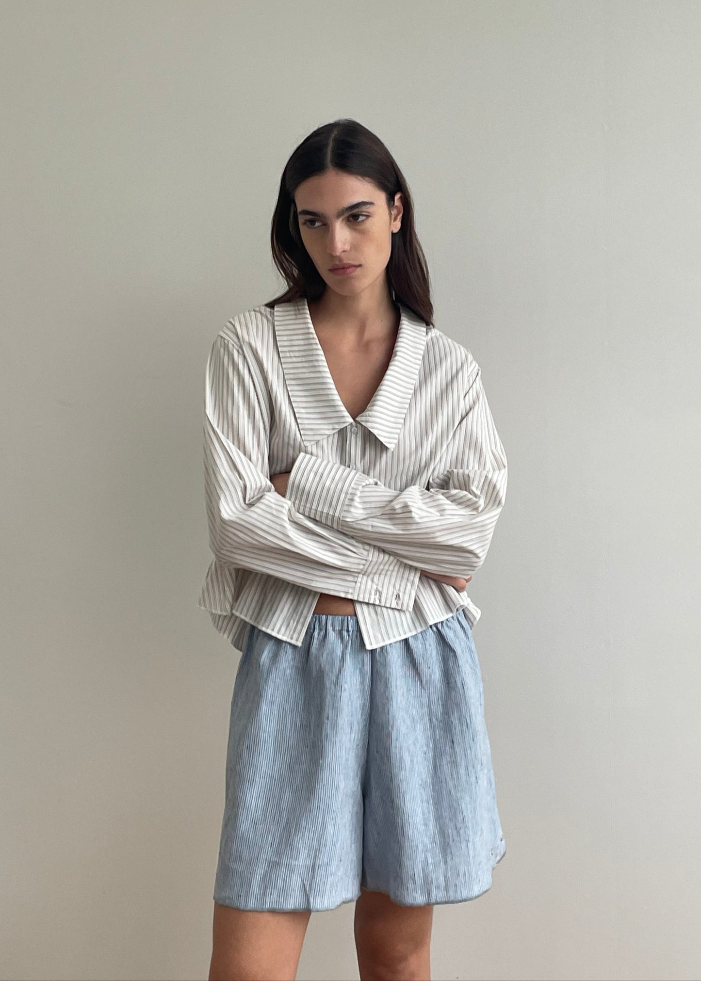 A woman with long dark hair stands against a light wall, arms crossed and expression neutral, wearing a loose Deiji Studios blue stripe long-sleeve shirt with a wide collar and Press Stud Short – Blue Stripe.