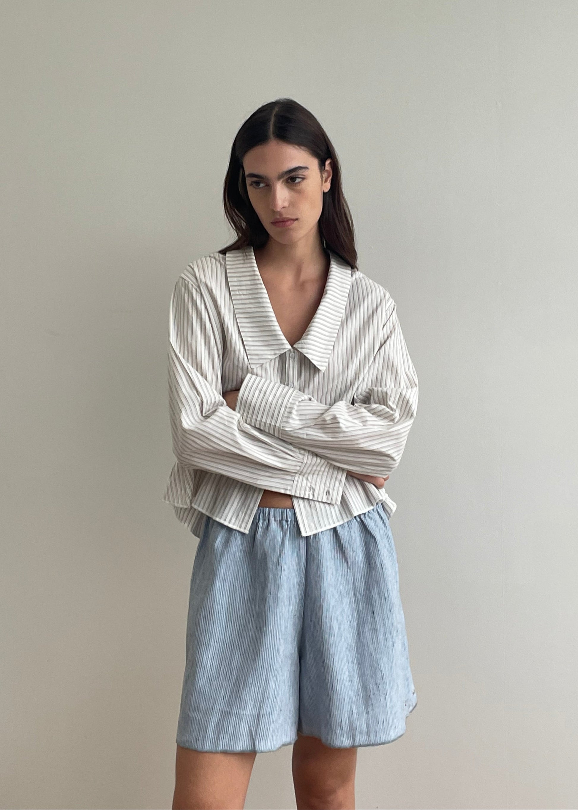 A woman with long dark hair stands against a light wall, arms crossed and expression neutral, wearing a loose Deiji Studios blue stripe long-sleeve shirt with a wide collar and Press Stud Short – Blue Stripe.