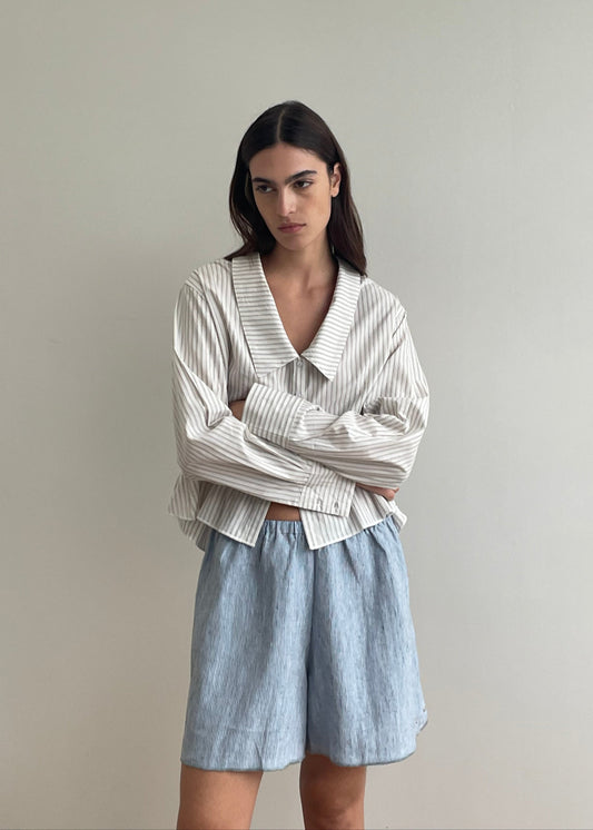 A woman with long dark hair stands against a light wall, arms crossed and expression neutral, wearing a loose Deiji Studios blue stripe long-sleeve shirt with a wide collar and Press Stud Short – Blue Stripe.