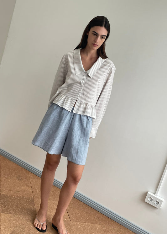 A woman stands indoors against a plain wall, barefoot, with straight dark hair, looking slightly downward. She wears a striped French linen long-sleeve blouse and the Press Stud Short - Blue Stripe from Deiji Studios.