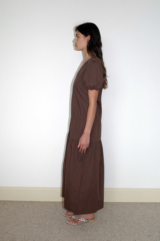A woman stands in profile against a plain white wall, wearing the Waistcord Dress - Carob Check by Deiji Studios. Her loose, wavy hair and relaxed pose complement the ankle-length silhouette and flat sandals for an effortlessly stylish look.