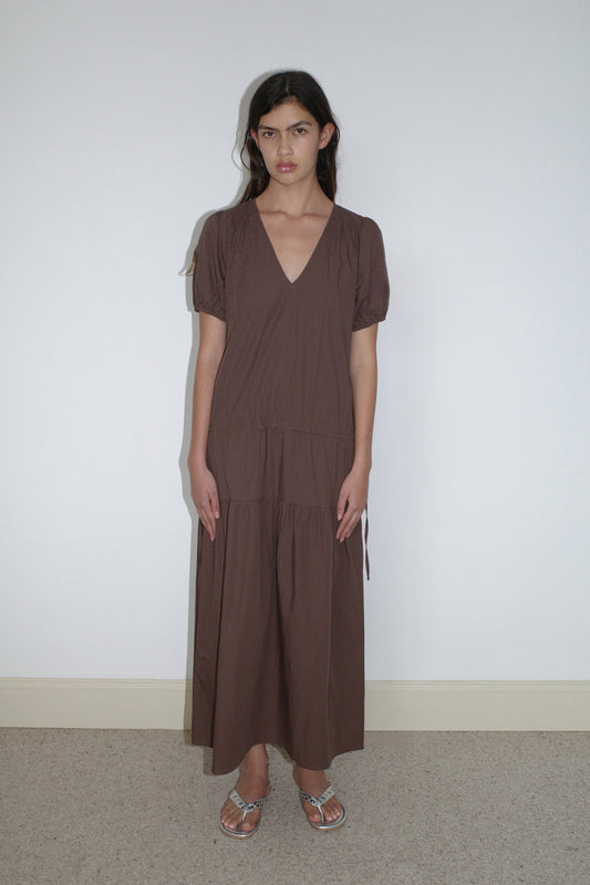 A woman with long dark hair and a neutral expression stands indoors against a plain white wall, wearing the Waistcord Dress - Carob Check by Deiji Studios with short sleeves and silver sandals.