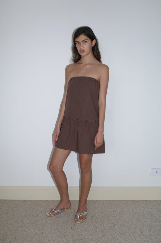 A woman stands indoors against a white wall, wearing the Button Dress - Carob Check from Deiji Studios and silver sandals. She has long dark hair, a neutral expression, and stands with one leg slightly bent.