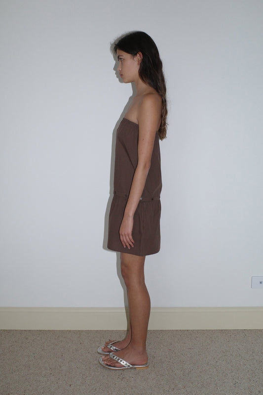 A young woman with long brown hair stands in profile against a white wall, wearing the Button Dress - Carob Check by Deiji Studios and white sandals on a beige carpeted floor.