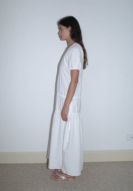 A woman with long brown hair stands in profile against a plain white wall, wearing the Waistcord Dress - White from Deiji Studios and sandals.