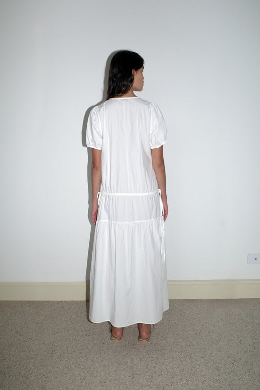 A woman stands with her back to the camera on a beige carpet, wearing the Waistcord Dress - White by Deiji Studios—an ankle-length, short-sleeved organic cotton dress—and beige heels against a white wall.