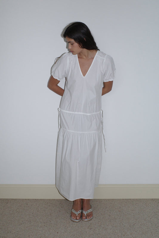 A woman stands against a plain white wall, looking down with her hands behind her back. She is wearing the Waistcord Dress - White by Deiji Studios, a loose-fitting, ankle-length organic cotton dress, paired with white sandals.