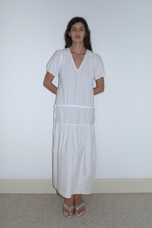 A woman stands against a plain white wall wearing the Waistcord Dress - White by Deiji Studios, an ankle-length organic cotton dress with short sleeves, V-neckline, and gathered details, paired with strappy flat sandals. Her hands are behind her back.