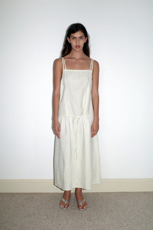 A woman with long dark hair stands against a plain white wall, wearing the Linen Strap Dress in Pastel Pistachio from Deiji Studios, featuring thin straps and a drawstring waist, paired with flat sandals.