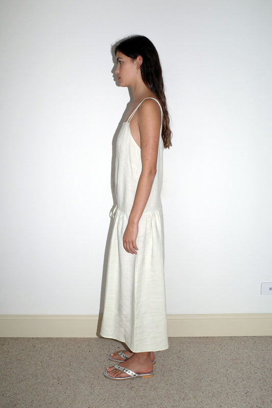 A woman stands in profile against a white wall, wearing the Linen Strap Dress - Pastel Pistachio by Deiji Studios and sandals. She has long, wavy brown hair and looks straight ahead.
