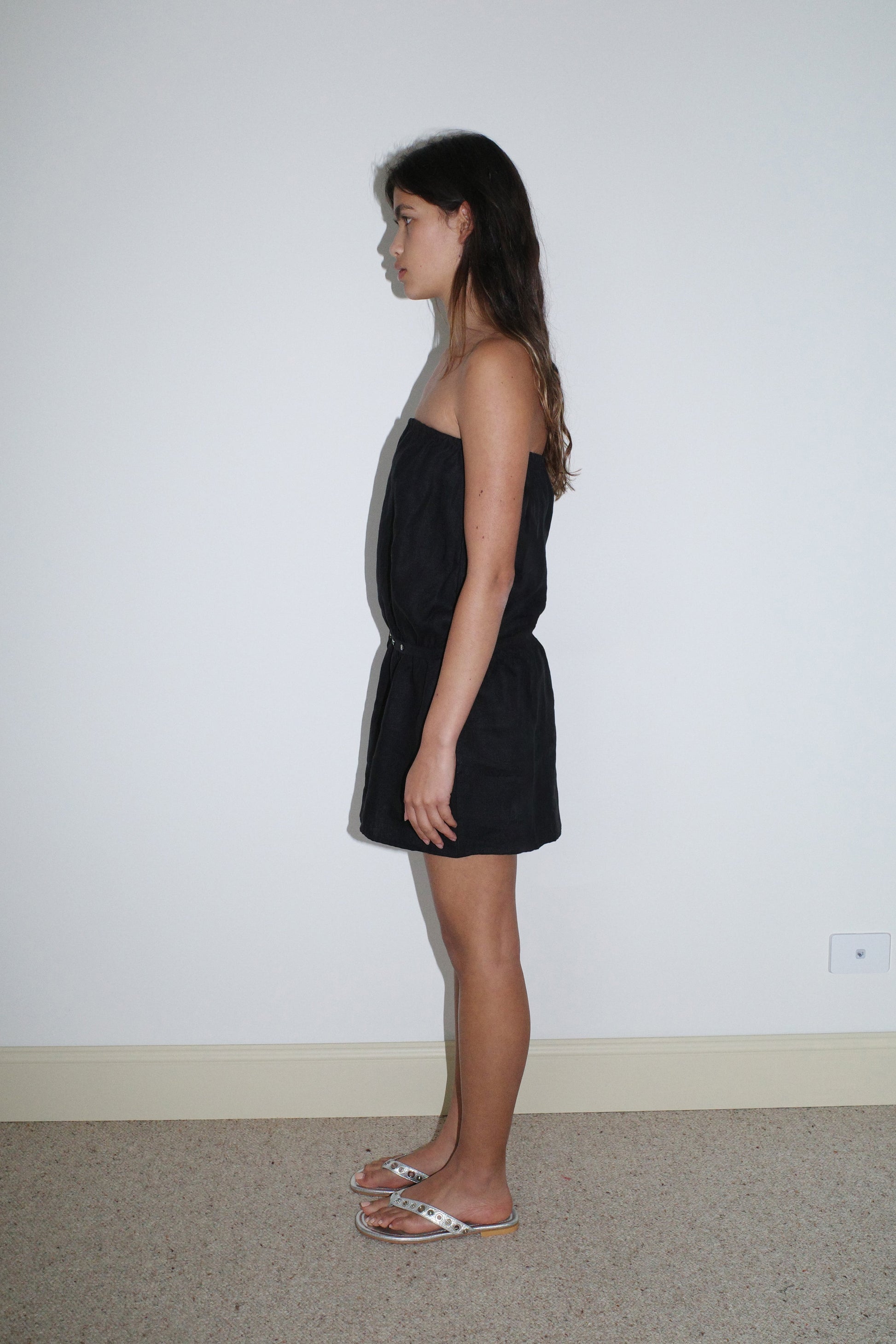 A woman with long brown hair wears the Strapless Linen Dress in black from Deiji Studios and white sandals, standing in profile against a plain white wall on a beige carpet.