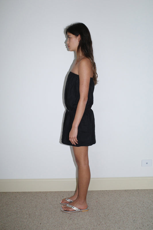 A woman with long brown hair wears the Strapless Linen Dress in black from Deiji Studios and white sandals, standing in profile against a plain white wall on a beige carpet.