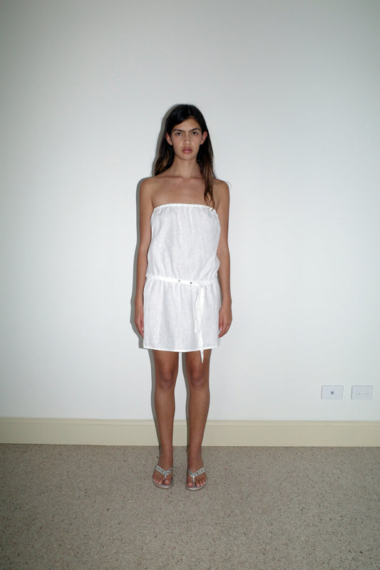A woman with long brown hair stands indoors against a plain white wall, wearing the Strapless Linen Dress - White. Her arms are relaxed at her sides, and the carpeted floor is visible.
