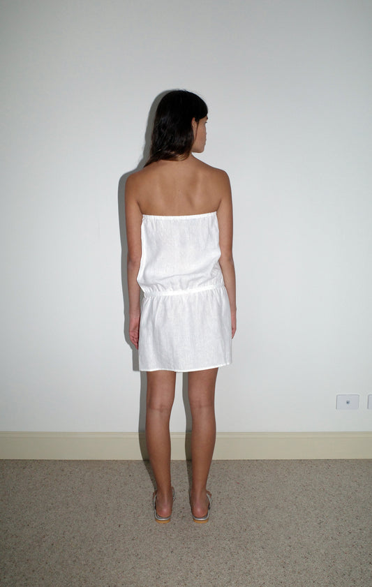 A woman with long dark hair stands on a beige carpet, facing a plain white wall. She wears the Strapless Linen Dress - White by Deiji Studios, made from organic French linen, and sandals. Seen from behind.