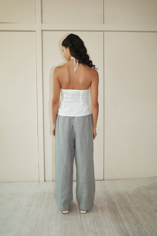 A woman with long dark hair stands facing away in a white halter top and the linen ease trouser - field check, featuring loose gray checked linen and an elastic waist, set against a minimalist light-toned indoor backdrop.