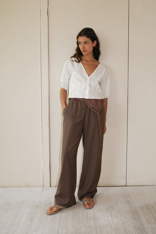A woman stands indoors against a beige wall wearing the Draw Top - White by Deiji Studios in organic cotton poplin, layered over a white blouse with brown pinstriped wide-leg pants and sandals, hands in her pockets as she gazes to the side.