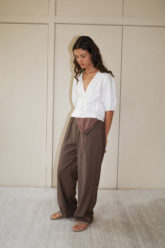 A woman with long curly hair stands indoors wearing the Draw Top - White by Deiji Studios and brown pinstripe wide-leg pants. She looks down calmly, standing against a beige wall.