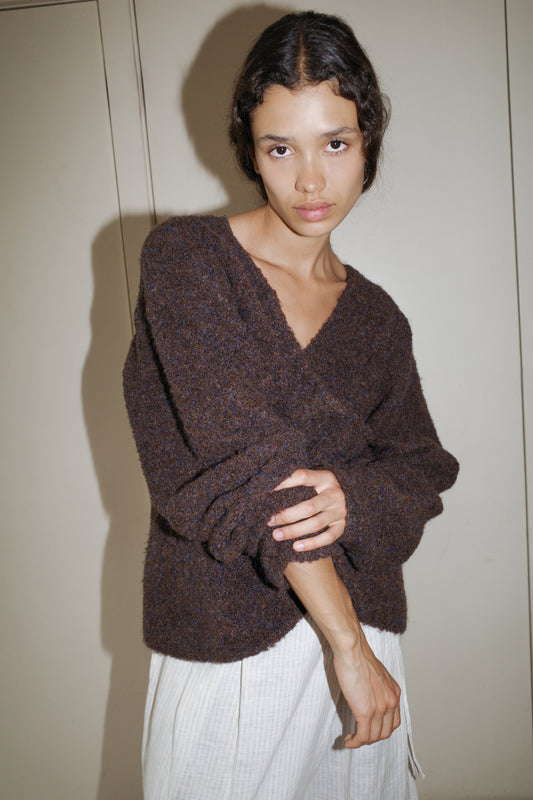 A person with wavy dark hair wears the Crossover Top - Brown Multi and light-colored pants, standing against a beige wall in a relaxed, slightly tilted pose.