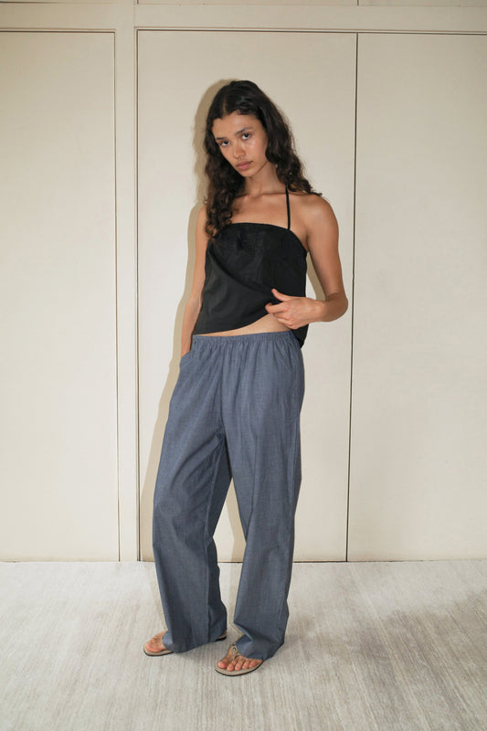 A woman with long curly hair stands indoors against a beige wall, wearing the Ease Trouser in Blue Pinstripe, paired with a black strapless top and sandals, her left hand resting on her top.