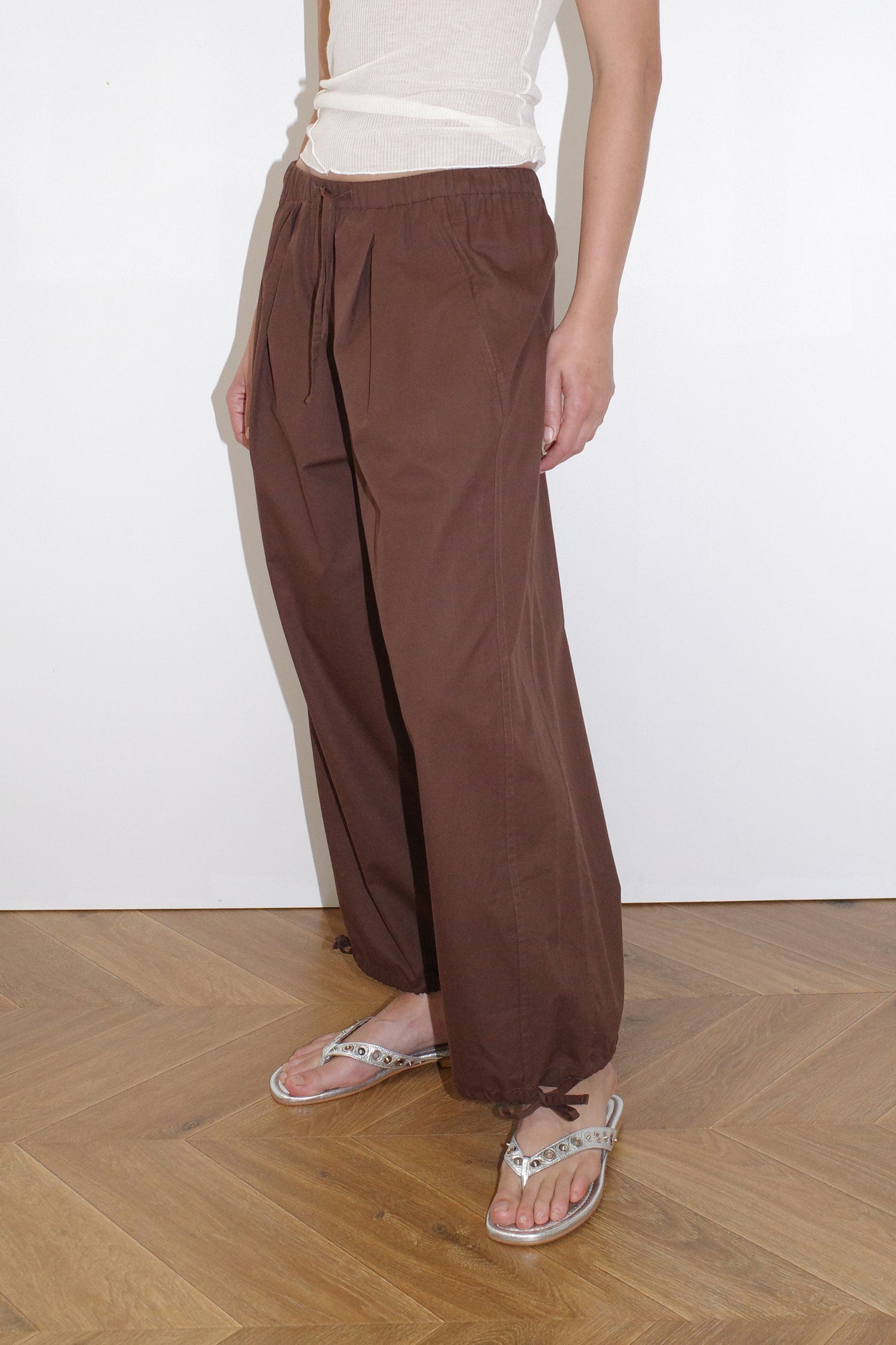 female model wears soft cotton trouser featuring a drawcord hem and waist with deep side pockets designed to sit mid-low waist for a relaxed look and made from 100% GOTS certified organic cotton. Paired with a knit tank in off white sheer fine knit tank with a low curved neckline and racer style back in a form fit, features an asymmetric baby lock exposed hem detail and finishes, made from a responsible blend of 65% ecovero viscose and 35% recycled polyester with OEKO-TEX 100 certification. 