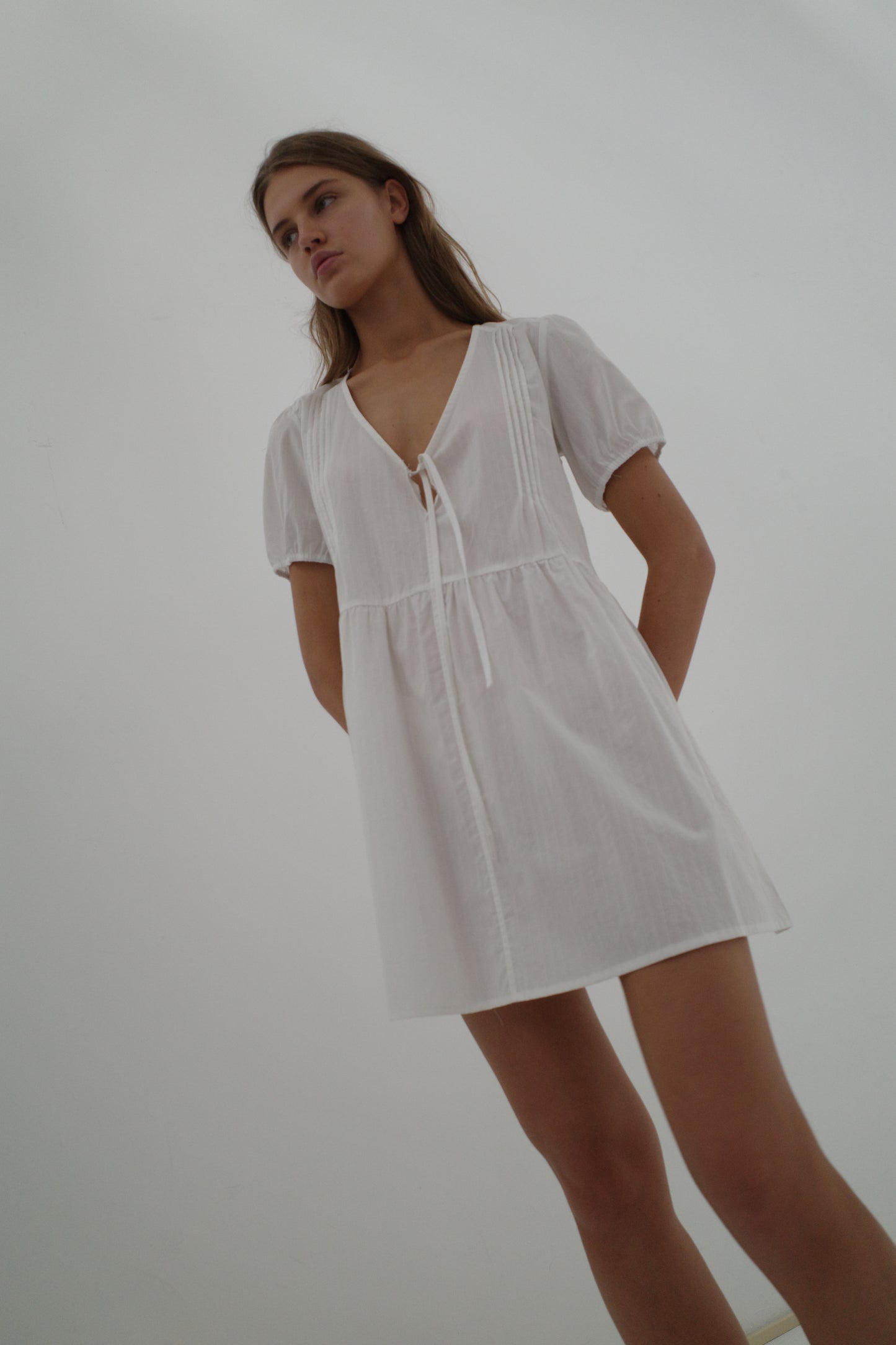 A young woman stands against a plain white background wearing the Deiji Studios V Pleat Dress - White Texture with short sleeves, a V-neck, and chest tie. She has long brown hair, gazing slightly to the side with her hands behind her back.