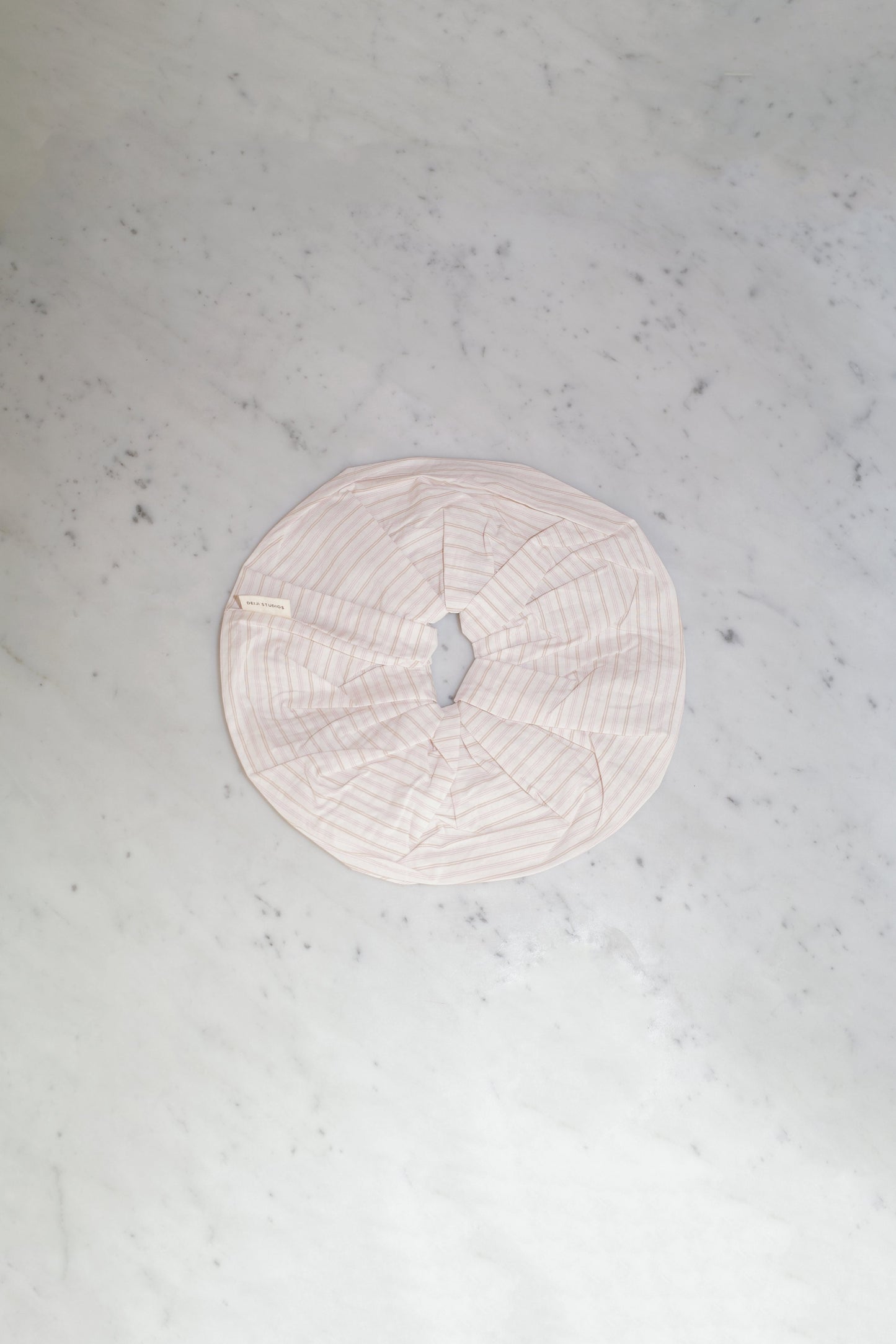 The Scrunchie - Dusty Stripe, an oversized round scrunchie in pale pink by Deiji Studios, made from padded organic cotton with a central opening, is displayed flat on a light gray marble surface.