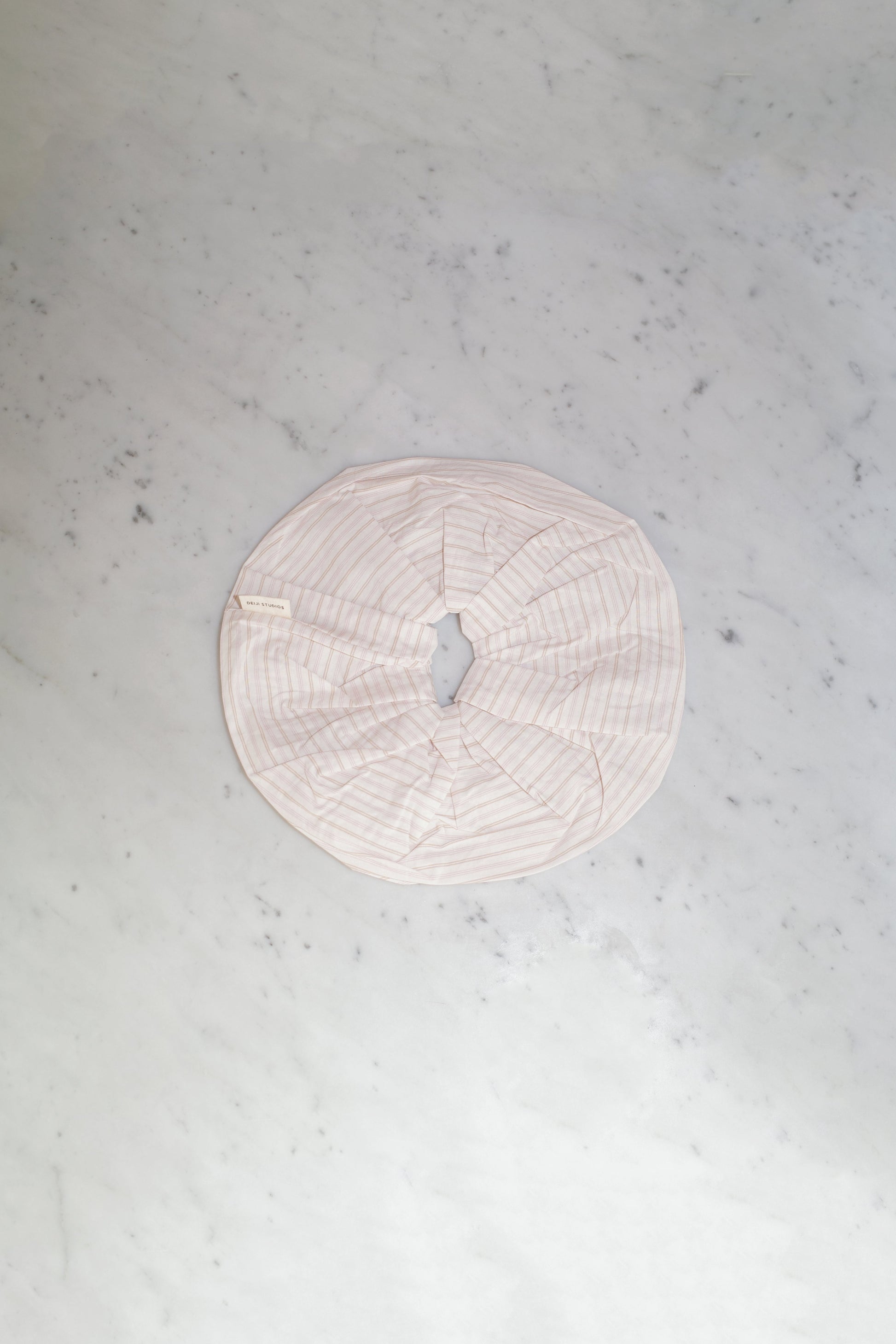 The Scrunchie - Dusty Stripe, an oversized round scrunchie in pale pink by Deiji Studios, made from padded organic cotton with a central opening, is displayed flat on a light gray marble surface.