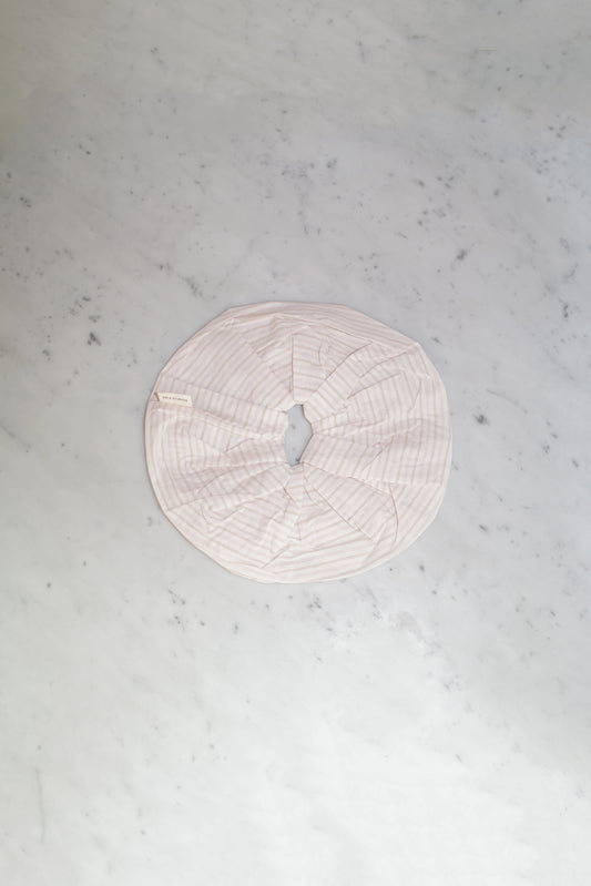 The Scrunchie - Dusty Stripe, an oversized round scrunchie in pale pink by Deiji Studios, made from padded organic cotton with a central opening, is displayed flat on a light gray marble surface.