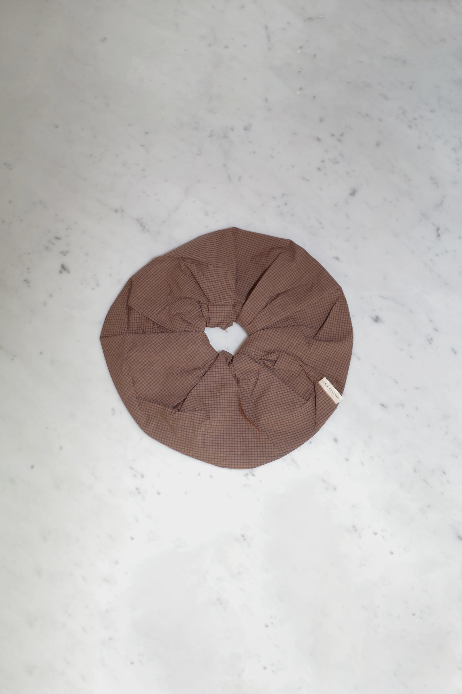 The Scrunchie - Carob Check, an oversized brown organic cotton scrunchie with a subtle checkered pattern from Deiji Studios, rests on a light grey marble surface.