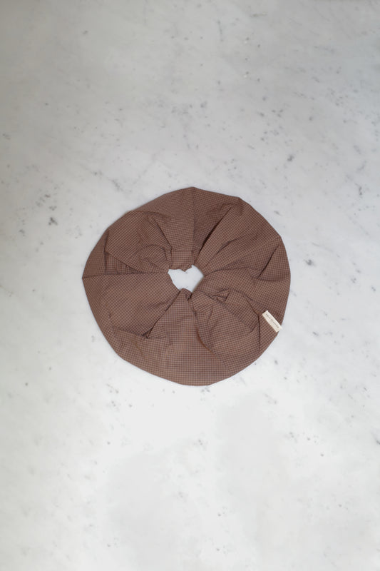 The Scrunchie - Carob Check, an oversized brown organic cotton scrunchie with a subtle checkered pattern from Deiji Studios, rests on a light grey marble surface.