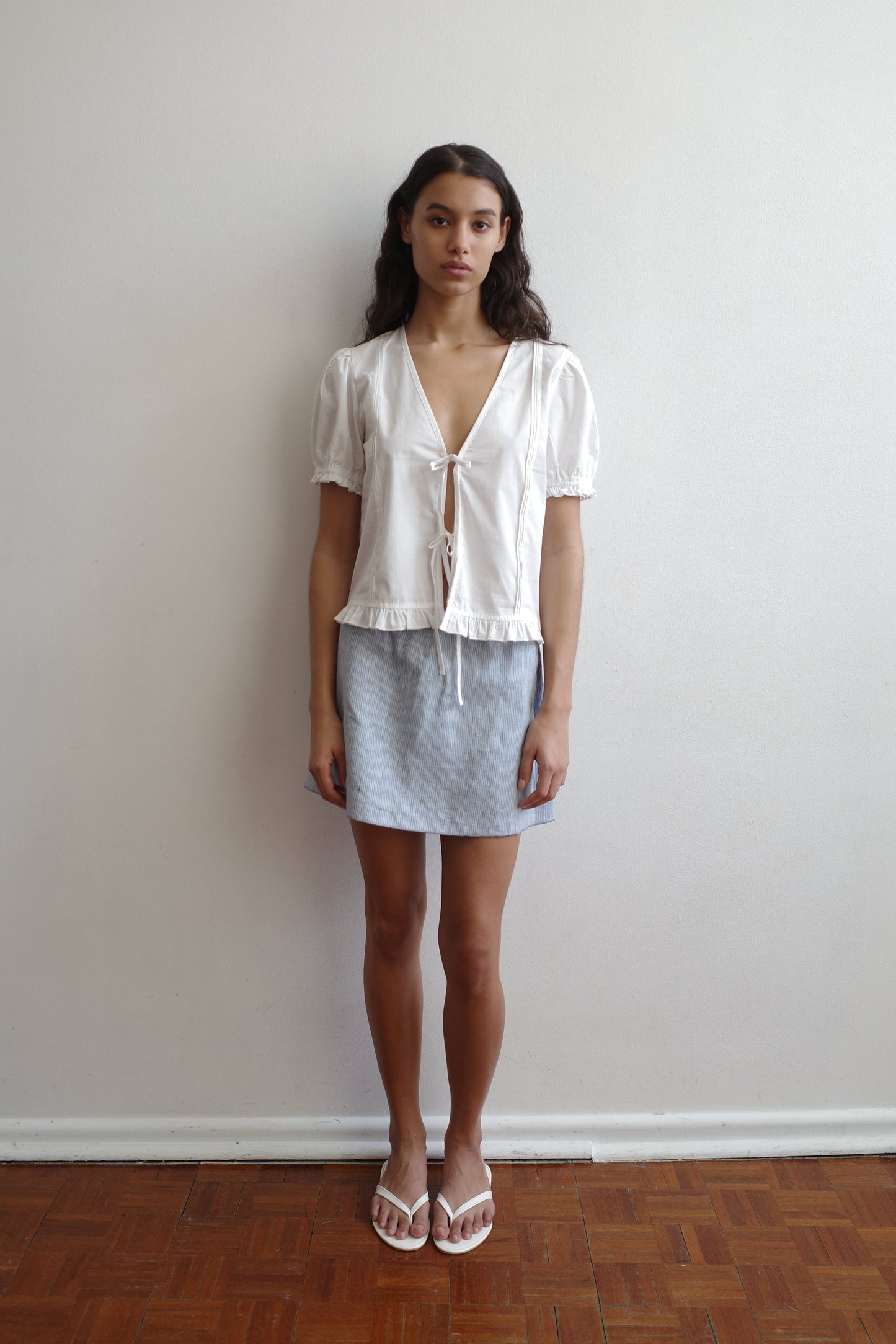A woman with long dark hair stands against a white wall in the Pintuck Top - White by Deiji Studios, paired with a light blue skirt and white sandals on a wooden floor.