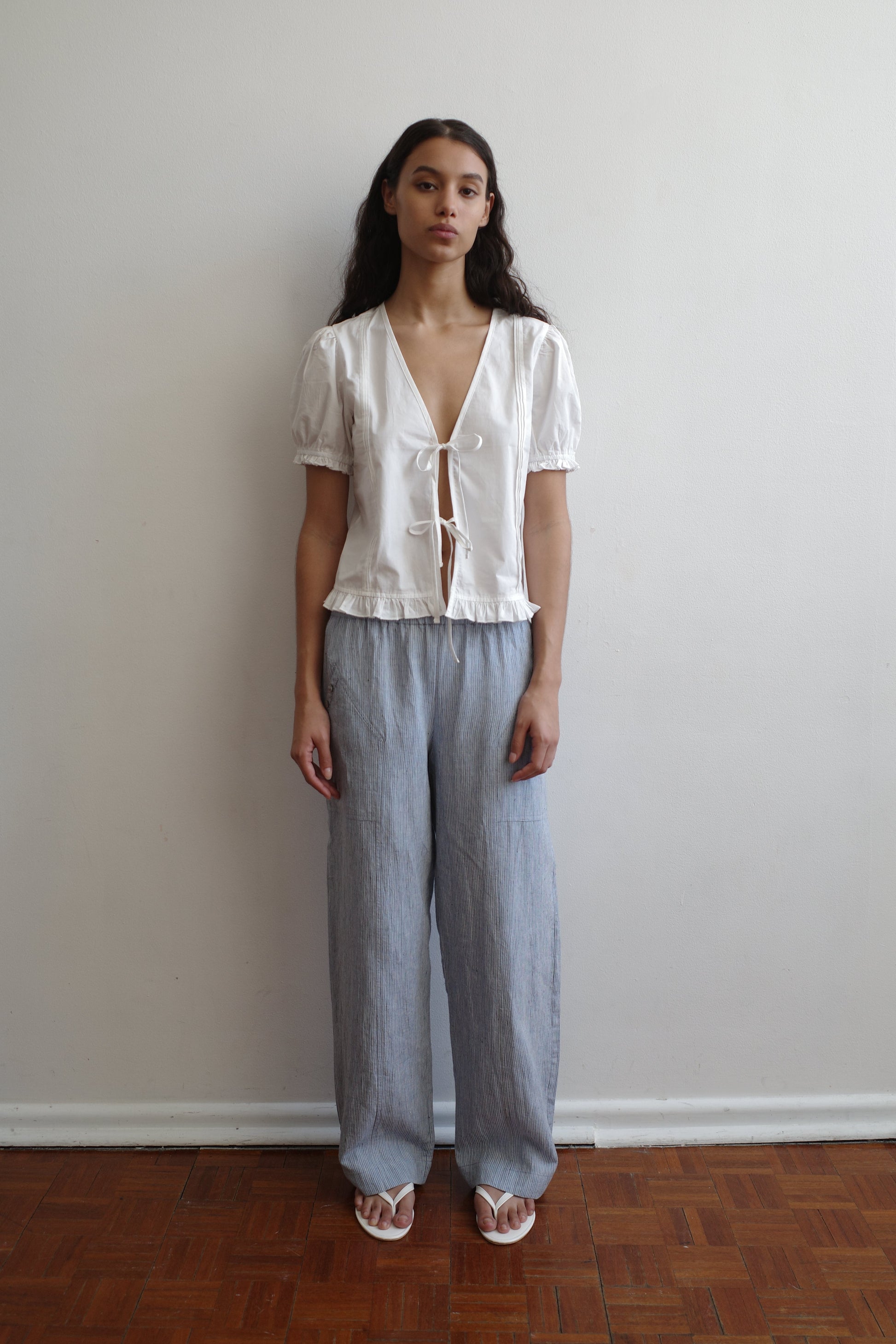 A woman with long dark hair stands indoors against a white wall, wearing a Pintuck Top - White by Deiji Studios featuring organic cotton poplin, pintucks, a deep V-neck with ties, light gray loose pants, and white sandals.