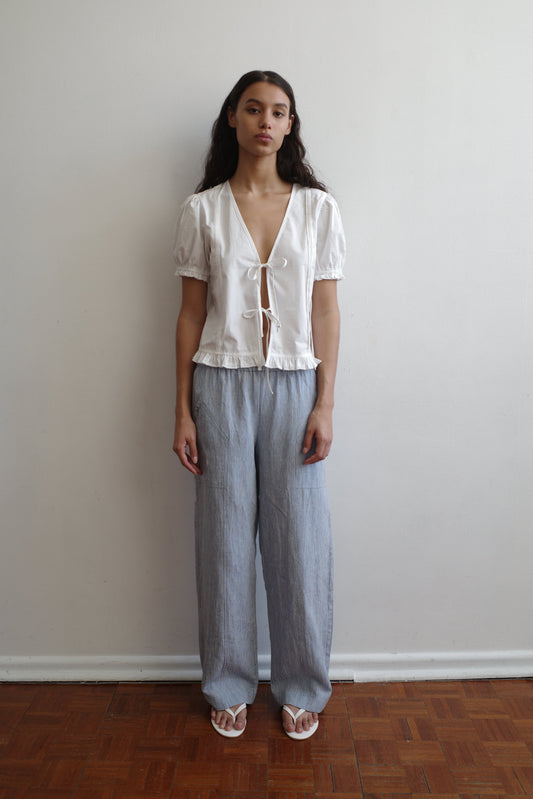 A woman with long dark hair stands indoors against a white wall, wearing a Pintuck Top - White by Deiji Studios featuring organic cotton poplin, pintucks, a deep V-neck with ties, light gray loose pants, and white sandals.