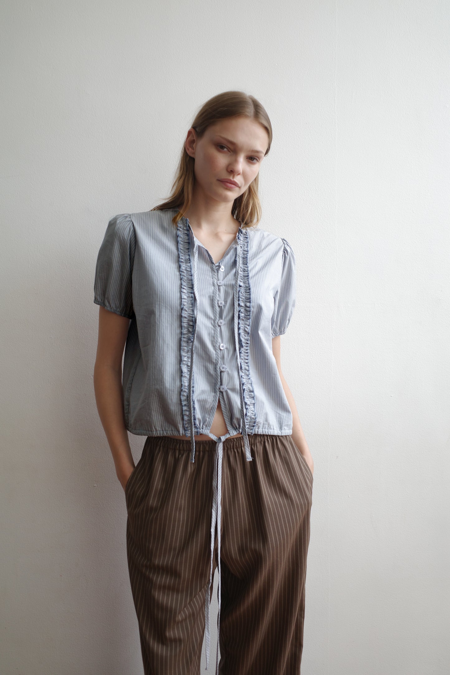 A woman with a neutral expression stands against a white wall, hands in her pockets, wearing the Double Frill Top - Sky Stripe by Deiji Studios and loose brown pinstriped pants.