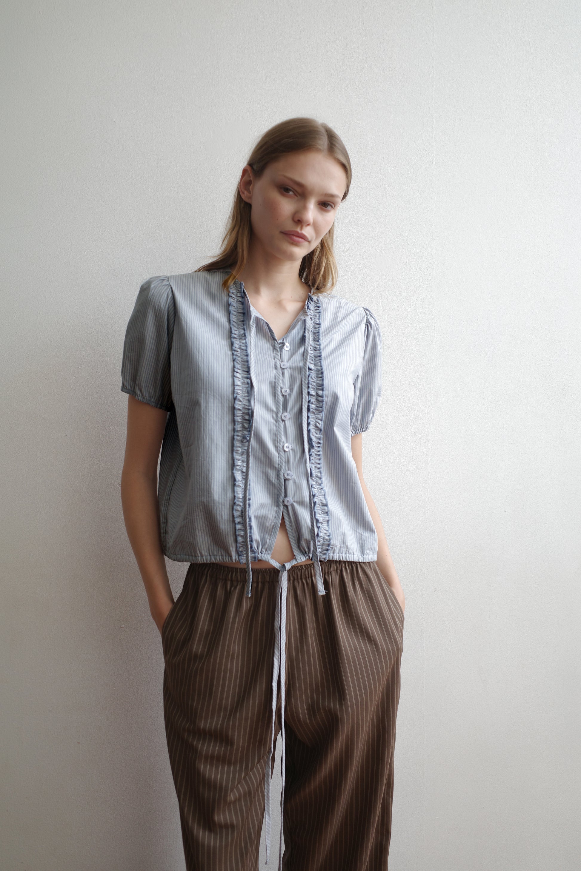A woman with a neutral expression stands against a white wall, hands in her pockets, wearing the Double Frill Top - Sky Stripe by Deiji Studios and loose brown pinstriped pants.