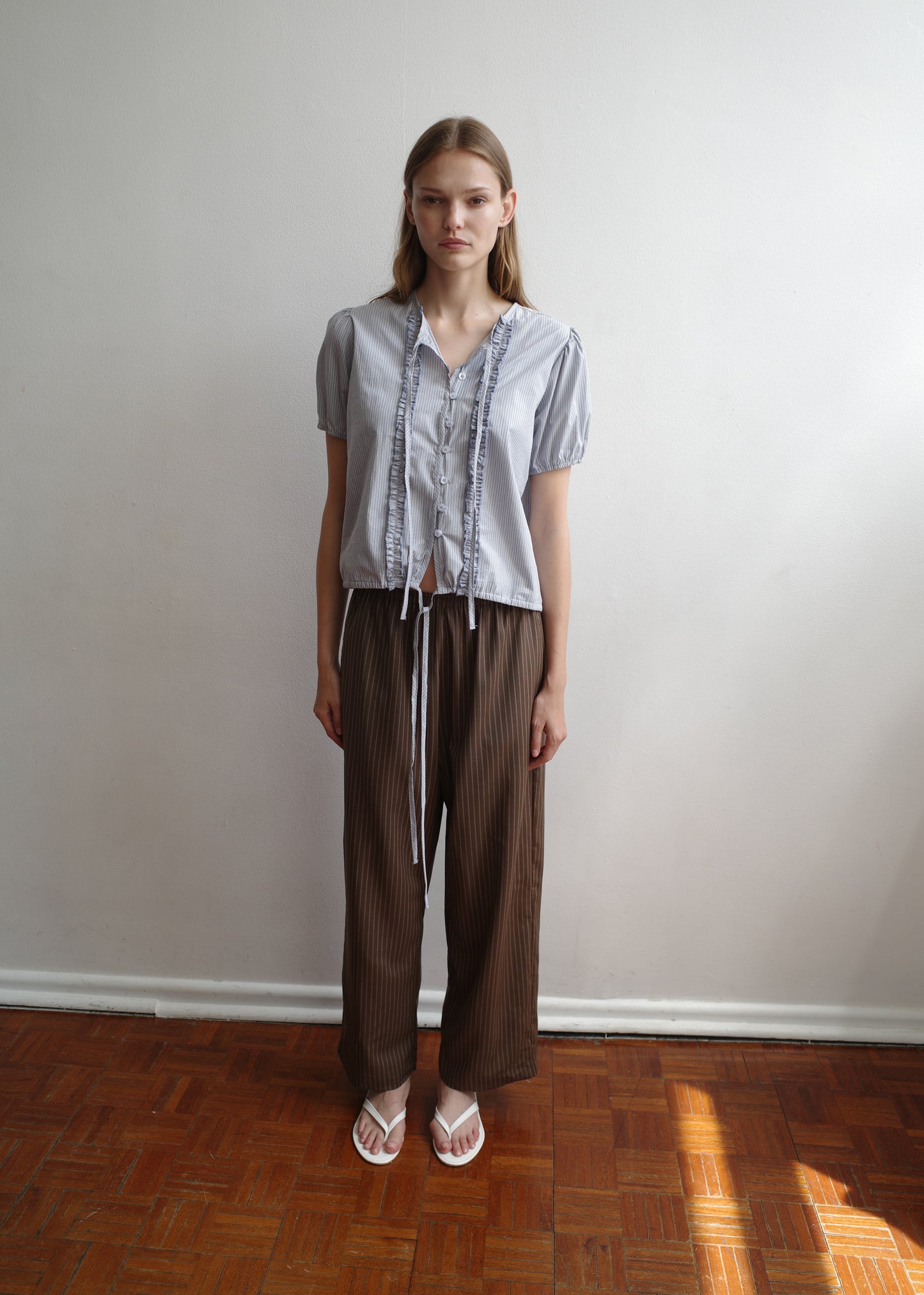 A woman stands indoors on a wooden floor, wearing the Double Frill Top - Sky Stripe by Deiji Studios with brown loose pants and white sandals, posed against a plain white wall.